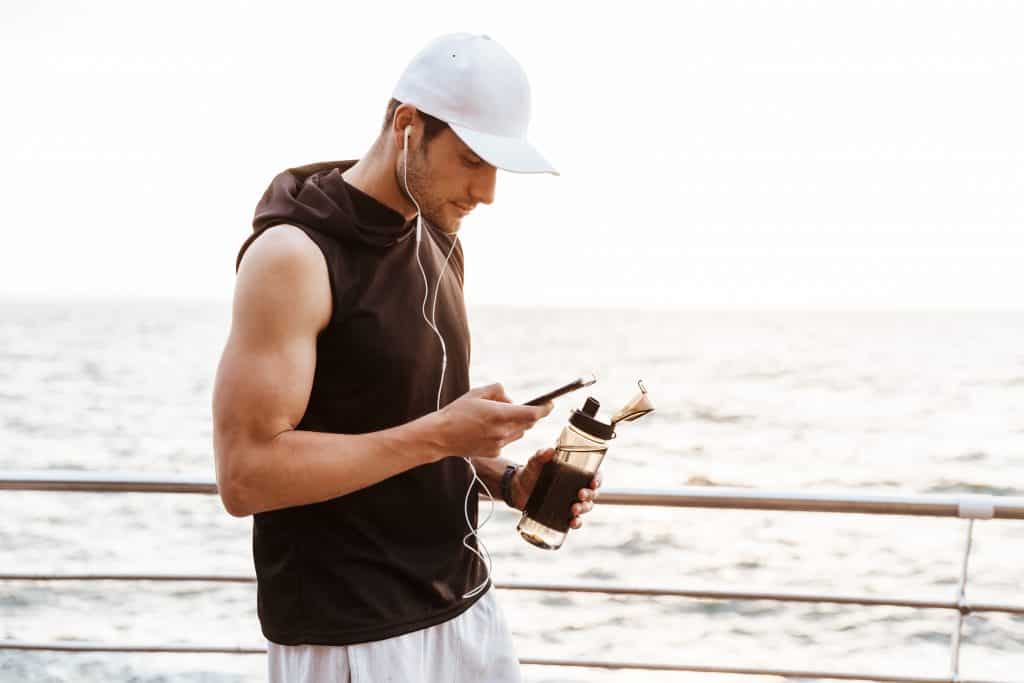 Photo closeup of handsome man in white cap using smartphone and drinking water while walking on pier at seaside in morning