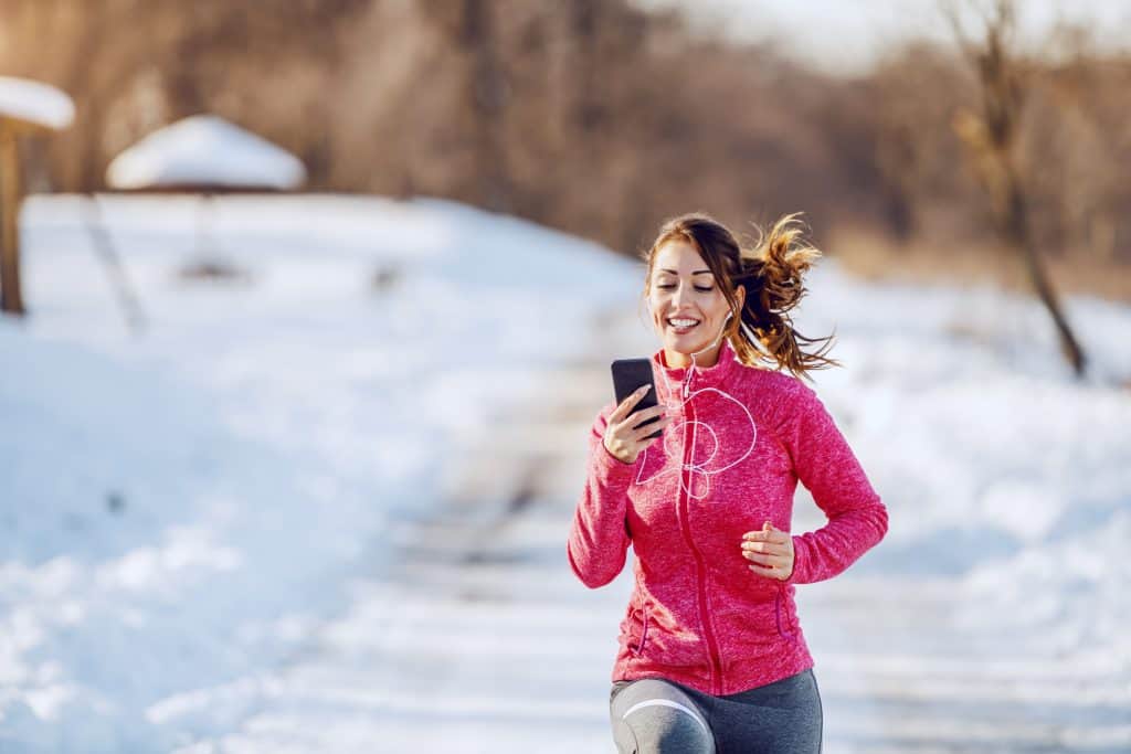 Smiling brunette in sportswear and with earphones in ears looking at smart phone while running in nature. Best Way to Carry Phone While Running