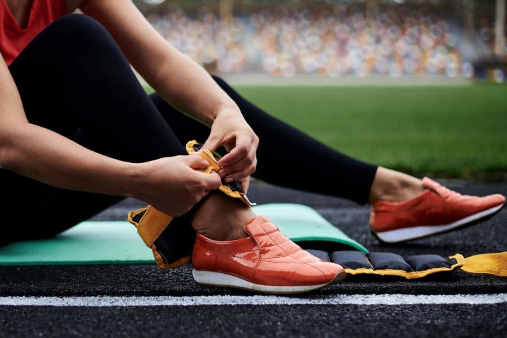 Close-up picture of woman's legs, showing process of putting on yellow ankle weights. Young woman, wearing black leggings and orange sneakers, is preparing herself for fitness training with weights.on