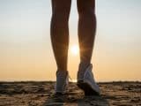Bottom view of woman legs in the sport sneakers on the beach at the morning opposite sea, rising sun, rays, beam