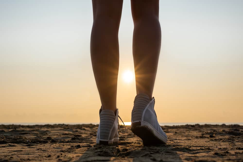 Bottom view of woman legs in the sport sneakers on the beach at the morning opposite sea, rising sun, rays, beam