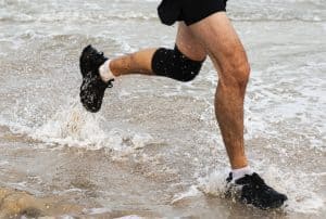 Man running with a knee brace on a beach: what benefit lies in wearing a knee brace for squats, how to pick the perfect one, and make the most out of it.