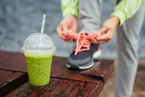 Green detox smoothie cup and woman lacing running shoes before workout on rainy day. Fitness and healthy lifestyle concept.