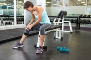 Fit brunette sitting on bench holding injured knee at the gym