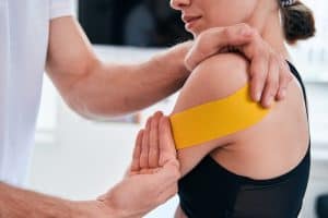Charming female patient sitting in medical office while professional kinesiologist applying yellow tape on her shoulder in medical center