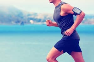 How Long Does It Take to Get in Shape Running: Cardio runner running listening smartphone music. Unrecognizable body jogging on ocean beach or waterfront working out with heart rate monitor app device and earphones in summer.