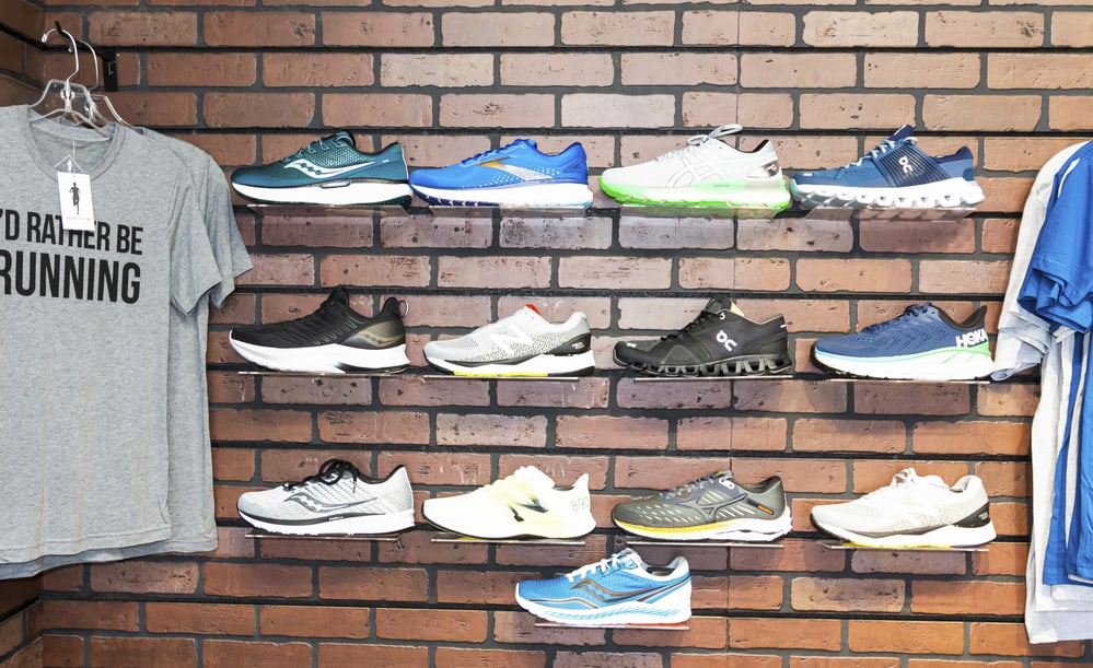 ASICS vs Nike Running Shoes: The wall of a running shoe specialty store has different brands of shoes displayed on the wall next to some t-shirts.